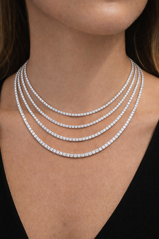 Close-up of a person wearing multiple diamond tennis necklaces on a black background showcasing a various of different diamond sizes